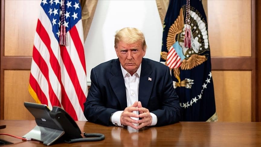 Donald Trump, başkanlık ofisinde masada otururken, arkasında Amerikan bayrakları ve başkanlık armasıyla birlikte.